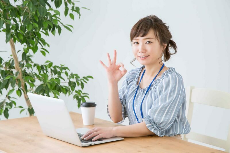 デスク上にあるパソコンを触りながら笑顔でokサインを出す20代の社会人女性